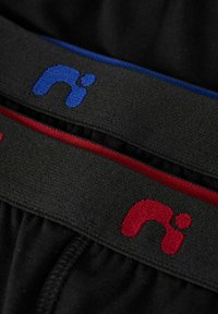 Cinturilla de tela negra con acentos de logotipo en rojo y azul. Las bandas presentan una textura suave y colores contrastantes sobre el material negro.