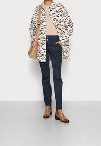 Koszulka w kremowym kolorze w wzory z czarnymi falistymi liniami, beżowa bluzka, granatowe spodnie i brązowe botki za kostkę. Prosta brązowa torebka na ramię.