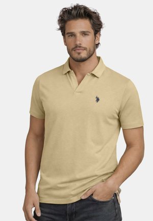 Mann mit kurzen braunen Haaren und Bart, der ein beiges Polohemd mit kleinem dunkelblauem Logo trägt, steht mit einer Hand in der Jeans-Tasche vor weißem Hintergrund.