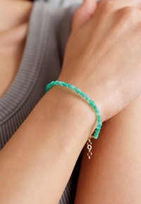 Bracciale di perline turchesi con chiusura in oro, caratterizzato da forme di pietre irregolari e un delicato accento di catena. Indossato su un polso abbronzato.