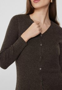 Cardigan marrón acanalado con escote en V, que presenta mangas largas y cinco botones pequeños en el frente. El material parece suave con un acabado texturizado.