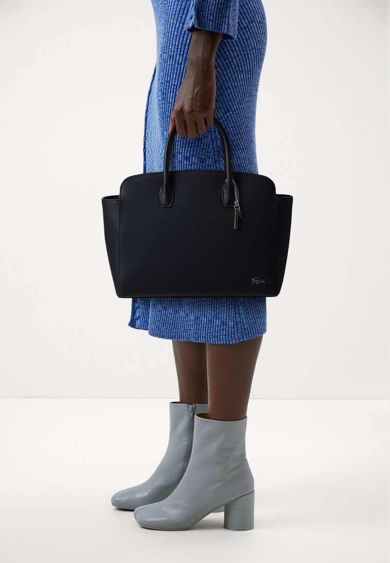 Borsa a tracolla in pelle nera con angoli arrotondati, manici superiori doppi e un charm laterale, abbinata a un vestito blu a coste e stivali grigi con tacco.