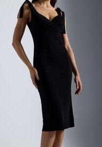 Robe noire ajustée en tissu doux. Comprend un décolleté profond en V, un détail torsadé sur le corsage et des bretelles nouées aux épaules. Longueur midi.