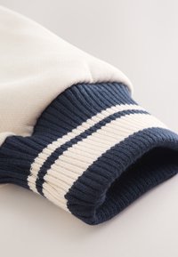 Gros plan d'une manche crème avec un poignet côtelé bleu marine comportant deux bandes horizontales crème.