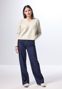 Pullover a costine color beige chiaro con colletto a V, abbinato a pantaloni in denim a gamba larga blu scuro e sneakers bianche. Insieme semplice e casual.