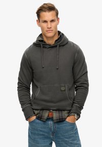 Dark gray hoodie with a front pocket, drawstring hood, and ribbed cuffs, paired with a plaid shirt and blue jeans. Casual fit.