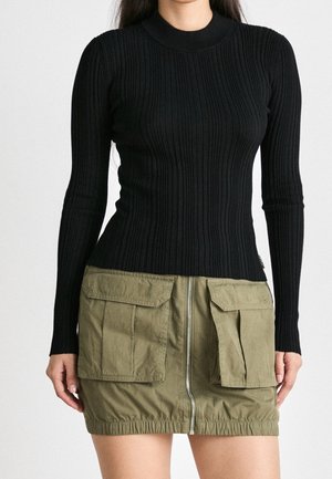 Kobieta ubrana w czarną prążkowaną bluzkę z długim rękawem oraz oliwkowo-zieloną mini spódniczkę typu cargo z przednim zamkiem i dużymi kieszeniami z klapami.