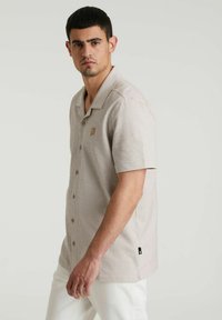 Camisa de botones de manga corta en tejido a rayas de color marrón claro y crema, con un logo en el pecho, cuello y detalle de etiqueta lateral.