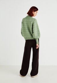 Green knitted sweater with a high neck, oversized sleeves, and textured finish, paired with dark, wide-leg ribbed pants.
