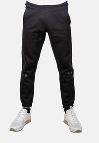 Pantaloni jogger neri slim-fit con polsini elastici indossati con sneakers bianche su uno sfondo chiaro e uniforme.