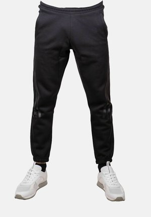 Pantalones jogger ajustados negros con puños elásticos, llevados con zapatillas blancas sobre un fondo claro y liso.