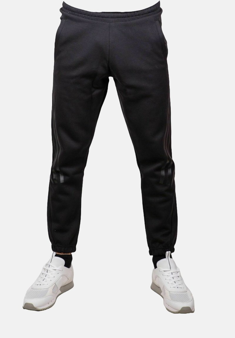 Pantaloni jogger neri slim-fit con polsini elastici indossati con sneakers bianche su uno sfondo chiaro e uniforme.