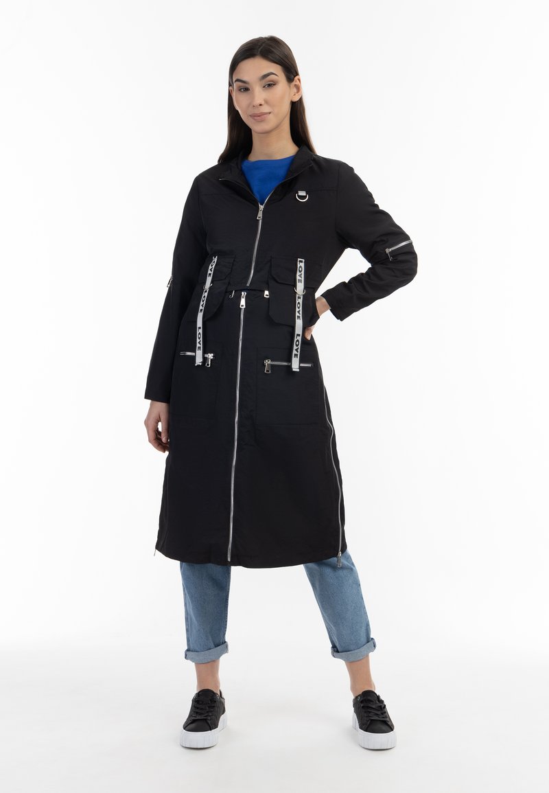 Giacca nera lunga con zip frontale, dotata di due tasche sul petto, cinture con testo "LOVE" e zip sui polsini delle maniche. Abbinata a jeans arrotolati e sneakers.
