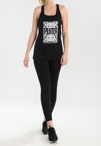 Black athletic tank top with a textured white PUMA logo design. Paired with black leggings and black sneakers. Smooth, fitted silhouette.