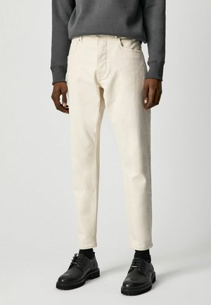 Mężczyzna ubrany w kremowe jeansy o prostych nogawkach, czarne skarpetki, czarne buty na sznurowadła i ciemnoszarą koszulę z długim rękawem, stojący na jednolitym tle.