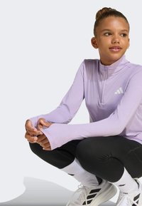 Light purple long-sleeve top with a quarter zipper, black leggings, and white athletic shoes. Features thumbholes and subtle branding.