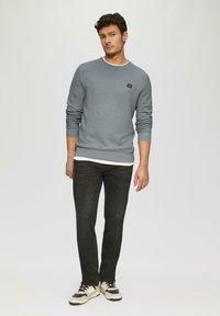 Grau strukturierter Pullover mit rundem Ausschnitt und gerippten Bündchen, über einem weißen Hemd getragen. Kombiniert mit schwarzen Jeans und sneakers mit beigen Akzenten.