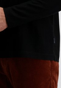 Zwarte longsleeve shirt met een visgraatstructuur, gecombineerd met roestkleurige corduroy broek. Het shirt heeft een zijlabel; de broek vertoont verticale ribbels.