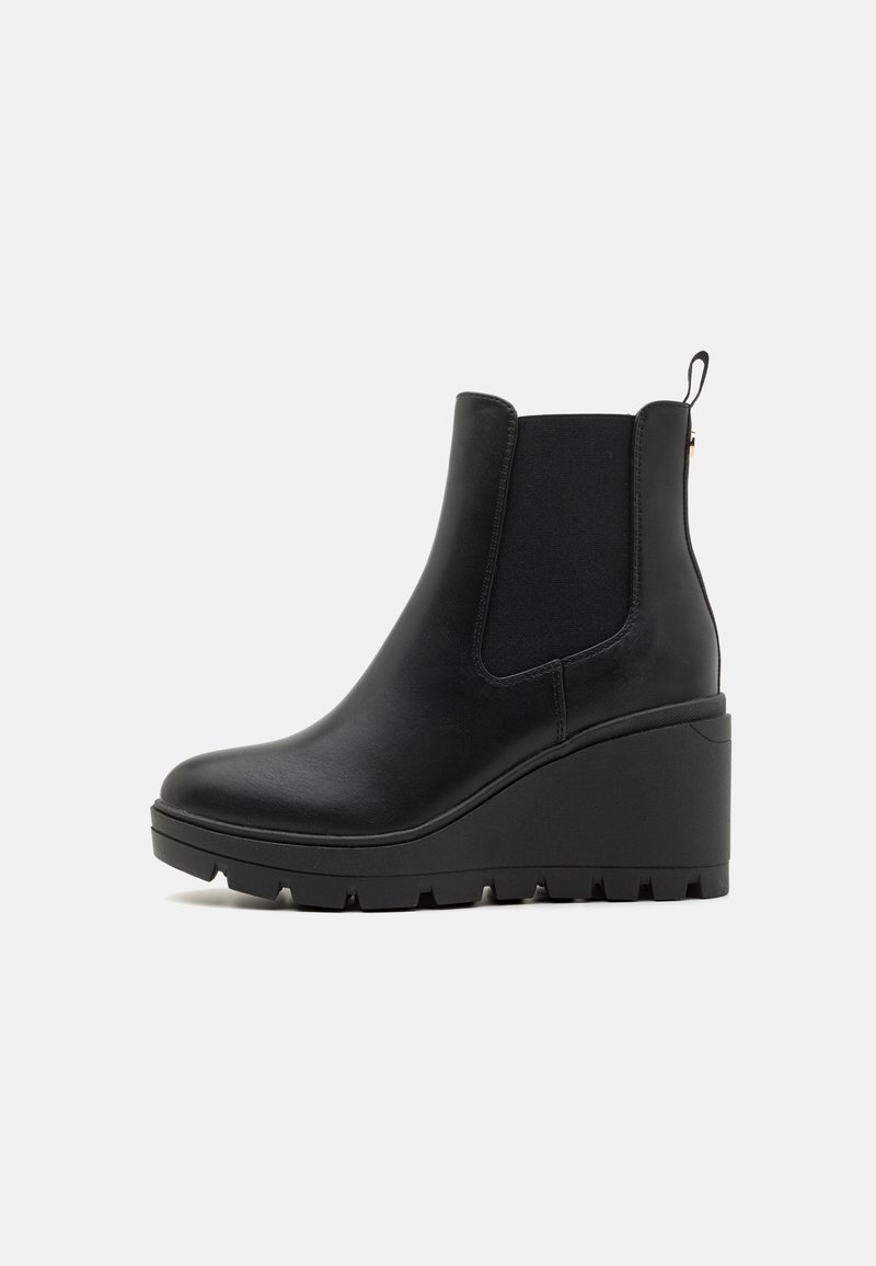 Bottine noire en cuir à talon compensé avec panneau latéral élastique et semelle rainurée sur fond blanc uni.