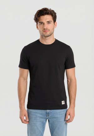 Hombre con cabello corto y castaño, vestido con una camiseta negra de manga corta lisa y jeans azul claro, de pie frente a un fondo claro.