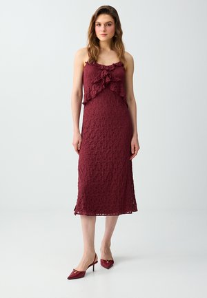 Frau in einem ärmellosen, burgunderfarbenen Spitzen-Midikleid mit Rüschen, kombiniert mit passenden spitzen Slingback-Pumps, stehend vor weißem Hintergrund.