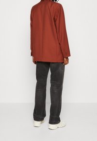 Personne portant un blazer couleur rouille, un jean noir délavé et des baskets blanches, debout de dos devant un fond blanc uni.