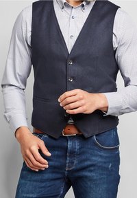 Mörkblå herringbone-väst med fem knappar, bärs över en ljus mönstrad skjorta. Har två framfickor och ett brunt bälte med en silverfärgad spänne.