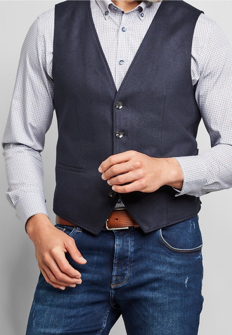 Mörkblå herringbone-väst med fem knappar, bärs över en ljus mönstrad skjorta. Har två framfickor och ett brunt bälte med en silverfärgad spänne.