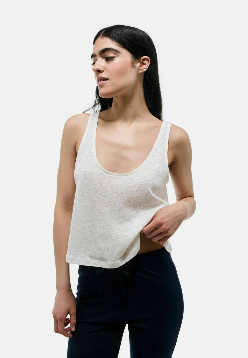 Crop top blanc sans manches en tissu léger, avec un décolleté à ras du cou et une finition légèrement texturée, associé à un pantalon foncé.