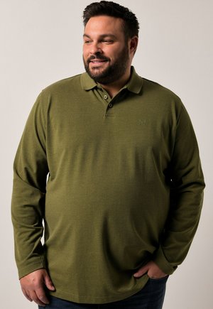 Hombre con barba que lleva una camiseta de polo de manga larga verde oliva, con una mano en el bolsillo, mirando ligeramente hacia un lado, fondo neutral.