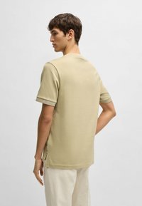 Camicia beige a maniche corte con tessuto texturizzato, scollo rotondo e spacchi laterali. Indossata sopra pantaloni larghi di colore chiaro.