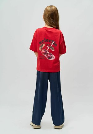 Camiseta corta roja con un gráfico juguetón de cereza y la palabra "ecléctico" en la parte de atrás, combinada con pantalones anchos de color azul marino.