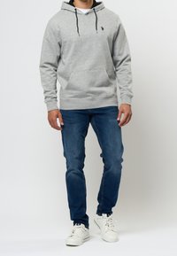 Felpa grigia con cappuccio, tasca a marsupio frontale, coulisse nere e piccolo logo; abbinata a jeans di denim blu e sneakers bianche.