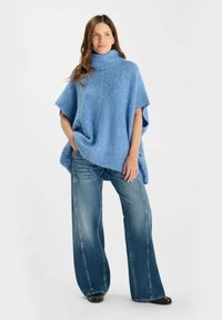 Poncho lavorato a maglia blu soffice con collo alto, abbinato a jeans a gamba larga blu chiaro e scarpe nere, su uno sfondo bianco semplice.