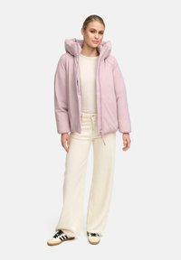 Vrouw in een lichtroze pufferjack over een beige trui, crèmekleurige wijde broek en witte sneakers met zwarte strepen.