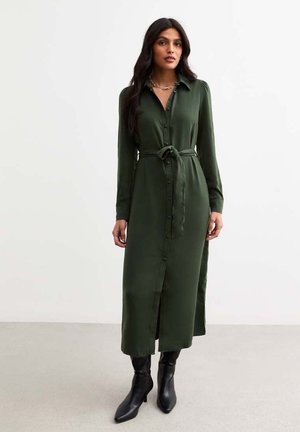Long green shirt dress made of smooth fabric, featuring a button-down front, collar, and a belted waist. Worn with black ankle boots.