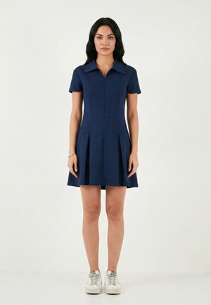 Naine seistes, kandes tumel sinist lühikeste varrukatega kraega kleiti voltidega, valgete tossudega, taustal hele ühtlane taust.