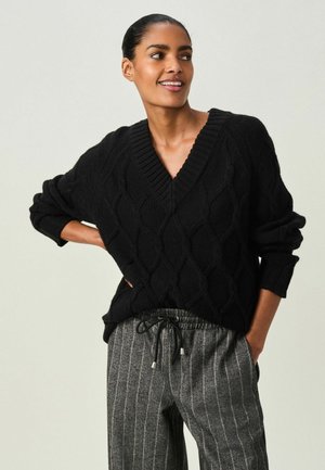 Maglia nera a maglia grossa con scollo a V e vestibilità comoda. Abbinata a pantaloni grigi a righe. Texture accogliente e dettagli del motivo sottili visibili.