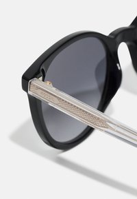 Lunettes de soleil noires avec une monture brillante et des verres gris. Branches transparentes avec un accent argenté texturé et un logo "FOSSIL".