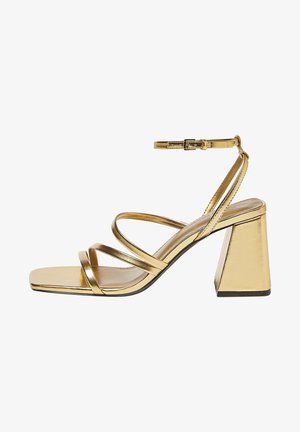 Sandal de tiras dorado con punta cuadrada, que presenta cuatro tiras metálicas y una correa ajustable en el tobillo, situada sobre un tacón de bloque robusto y angulado.