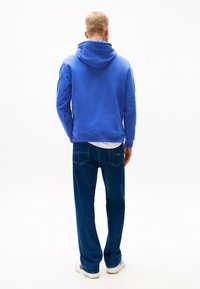 Blauer Hoodie mit strukturierter Oberfläche, lockerer Passform und großer Kapuze. Kombiniert mit dunkelblauen Jeans, die ein lässiges Outfit von hinten zeigen.