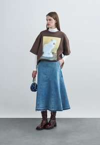 Bruin oversized T-shirt met witte kattenafbeelding, gecombineerd met een denim rok met uitlopende zoom. Model draagt kniehoge bordeauxrode laarzen en houdt een tas vast.