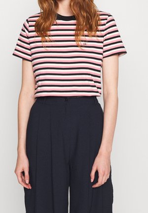 Mujer con una camiseta de manga corta a rayas en rosa, blanco y negro, metida en unos pantalones negros de talle alto, de pie frente a un fondo liso.