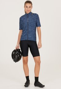 Kortärmad blå cykeltröja med dragkedja, i kombination med svarta vadderade shorts. Svarta cykelskor och en svart hjälm hålls i vänster hand.