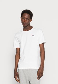 Camiseta de algodón blanca con cuello redondo, mangas cortas y un pequeño logotipo de FILA en rojo y azul en el pecho. Textura suave y lisa.