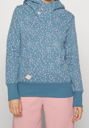 Osoba ubrana w niebieską wzorzystą bluzę z kapturem z różowymi i białymi kształtami liści oraz różowe dresy Adidas, stojąca na tle jednolitego białego tła.