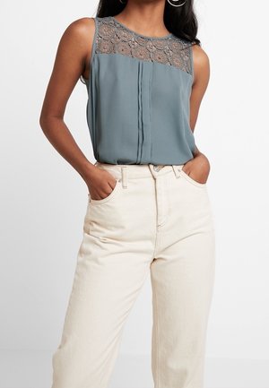 Femme portant un chemisier bleu sans manches avec un empiècement en dentelle, rentré dans un jean écru taille haute, mains dans les poches.