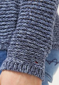 Gros plan sur la manche d'un pull bleu tricoté avec un motif texturé, portée sur un jean en denim bleu clair avec un cœur brodé.