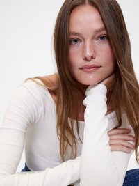 Biela rebrovaná blúzka s dlhým rukávom a širokým výstrihom. Modelka má dlhé, rovné hnedé vlasy a opiera si bradu o ruku.