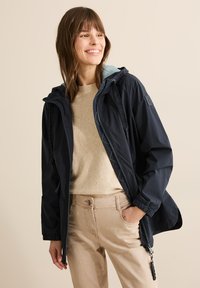 Chaqueta impermeable de color azul marino con cremallera frontal, capucha ajustable, forro contrastante en azul claro y puños elásticos, combinada con un suéter beige y pantalones.